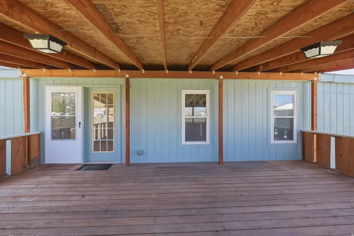 Expansive covered wooden deck featuring exposed wood beams, two exterior light fixtures, and a light blue siding backdrop