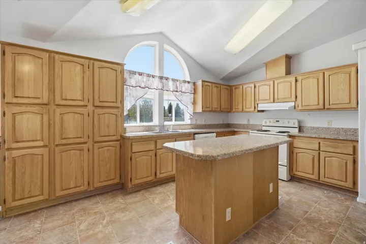Spacious kitchen featuring light wood cabinetry, a central island with a stone-finish countertop, and a vaulted ceiling with an arched window