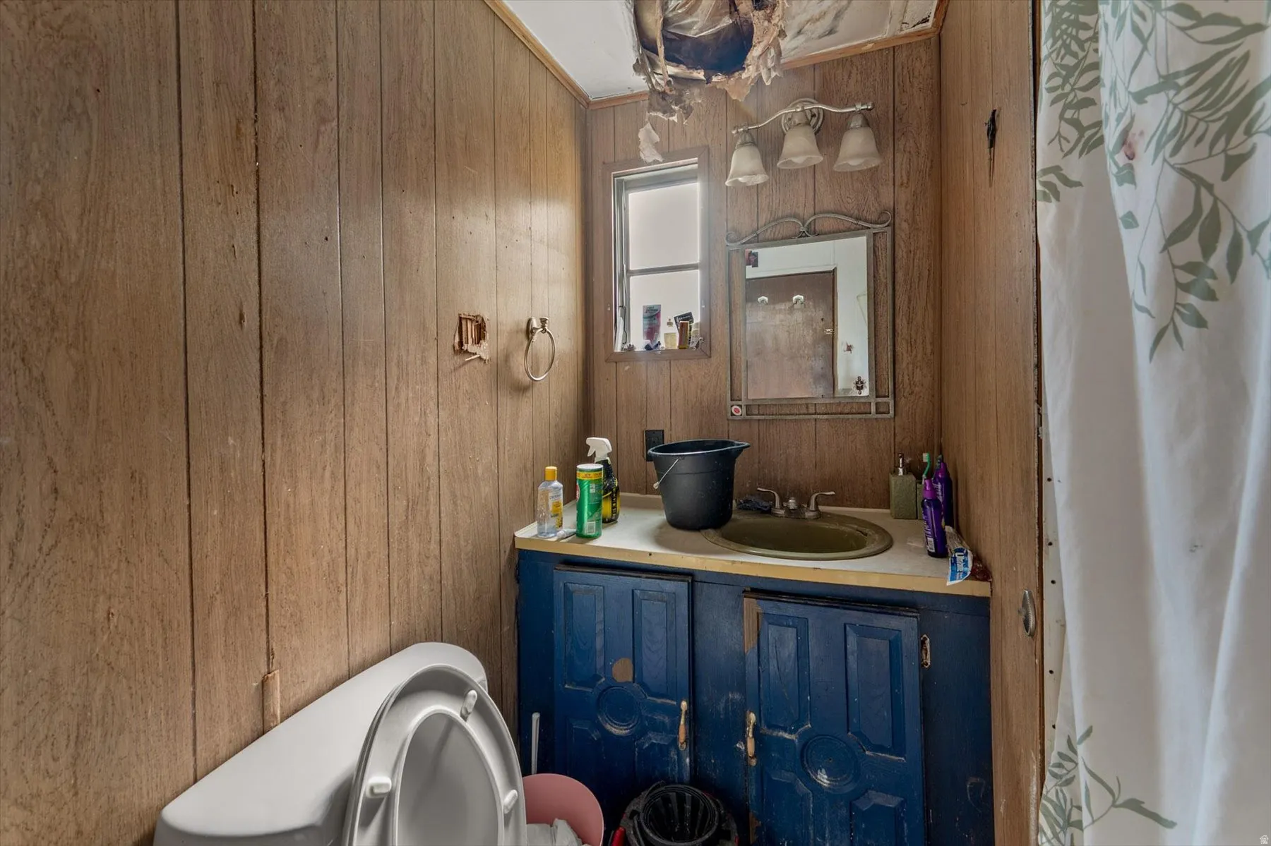 Bathroom featuring wood-finish paneling, a single window, vanity with painted blue cabinetry, and a white toilet
