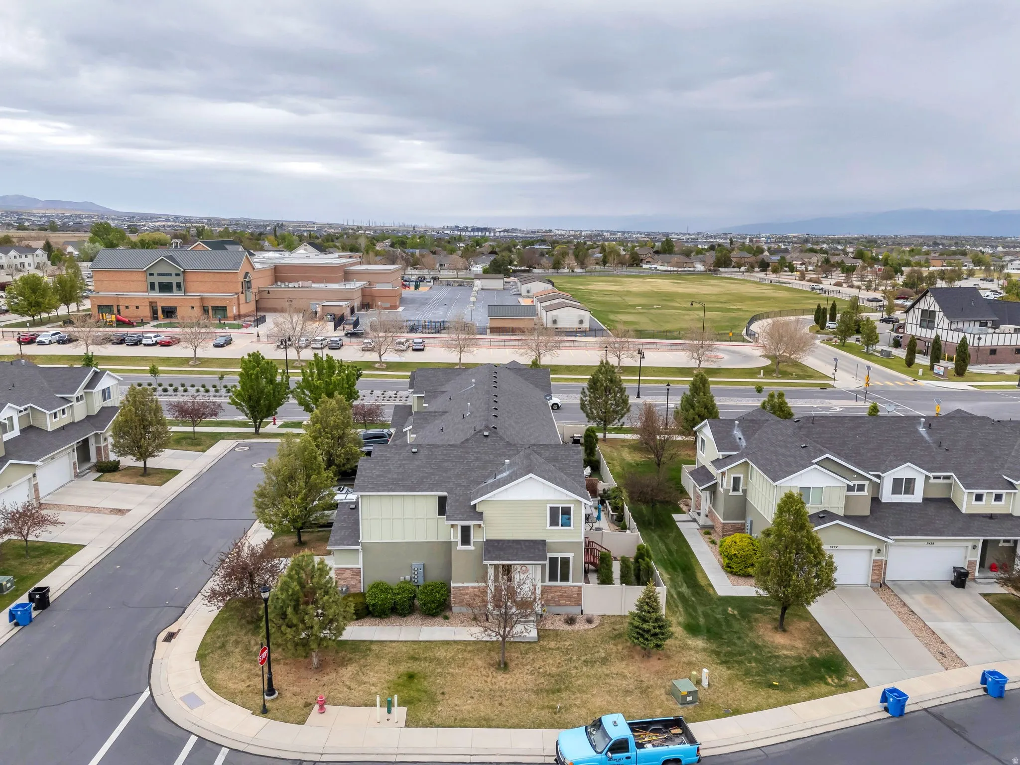 Aerial perspective showcasing a residential community with multi-story homes, a large institutional building, open fields, and mountain range backdrop