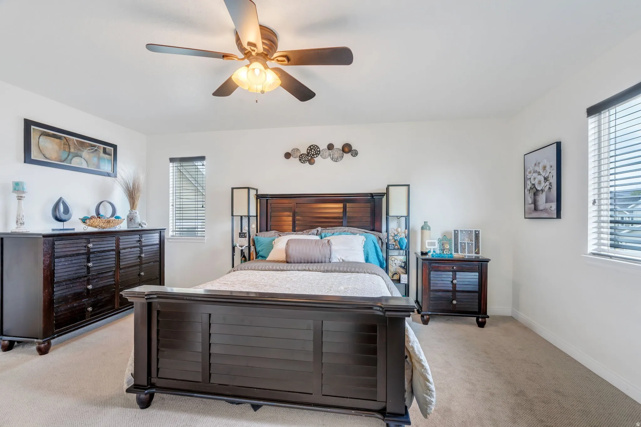 Bright bedroom featuring a ceiling fan, dual windows with blinds, light-colored carpeting, and white walls