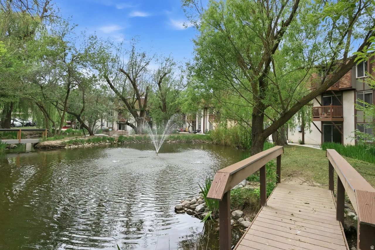 Expansive pond featuring a central fountain, bordered by mature trees and a pedestrian bridge with wood-finish decking
