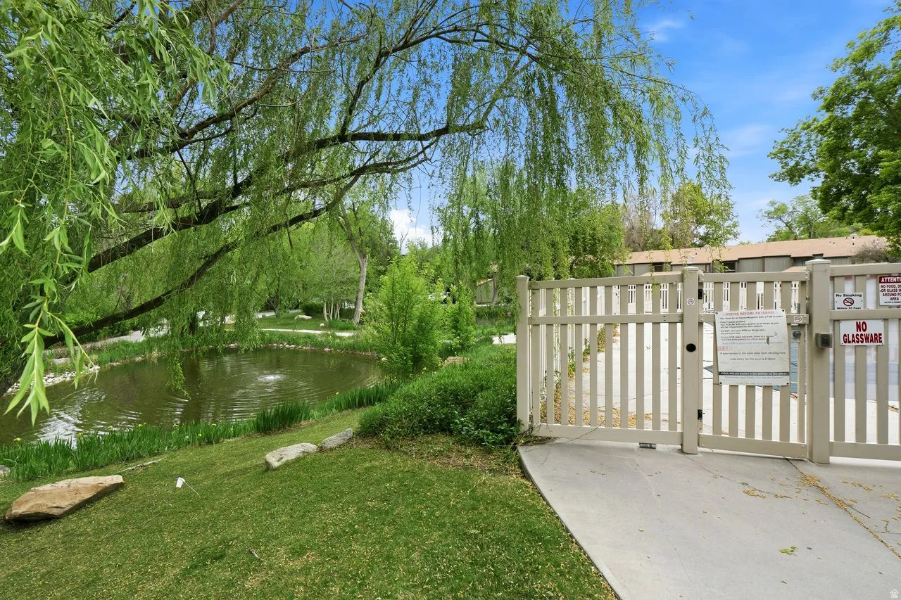 Lush landscaping featuring a serene pond with a water feature, mature trees, green lawn, and a light-colored slatted fence with access gates