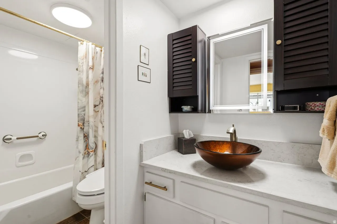 Bathroom featuring a vessel sink with a bronze-finish faucet, an integrated countertop, white cabinetry with gold-tone hardware, a new modern mirror with dark wood-finish cabinets.