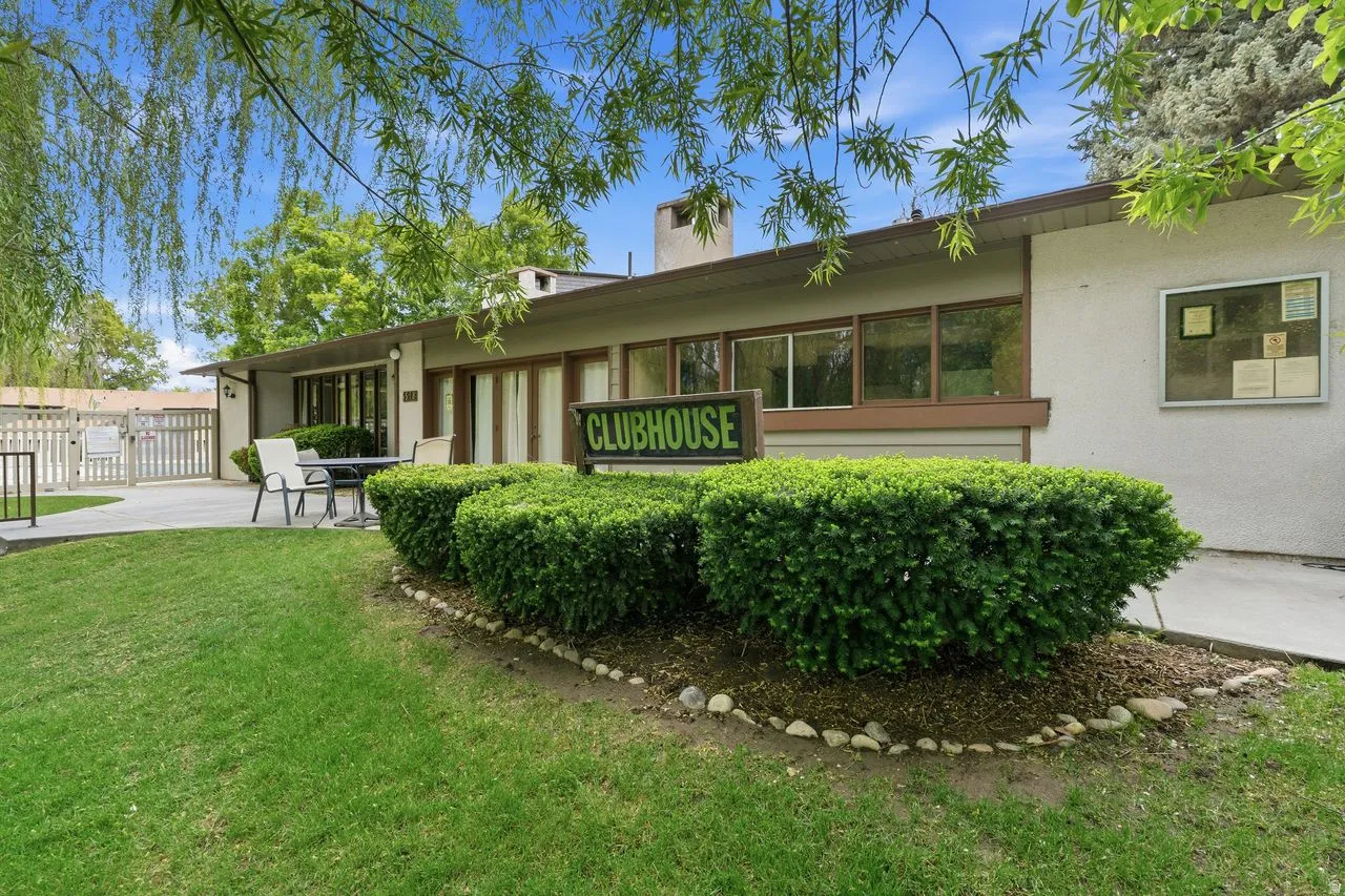 Clubhouse exterior featuring a neutral facade, multiple windows, a concrete patio, a white picket fence, and a green lawn with established landscaping