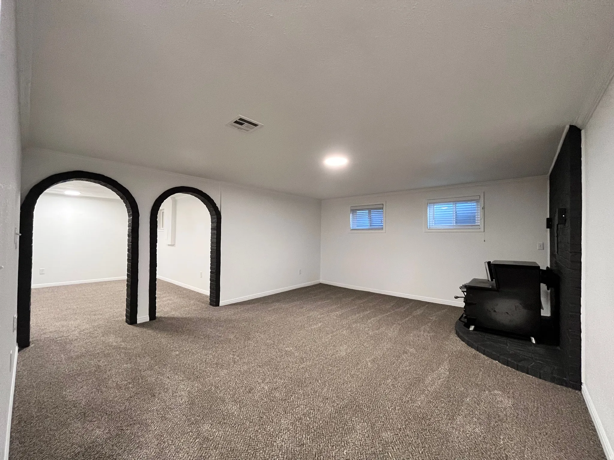 Spacious carpeted room featuring a corner-set wood-burning stove with a brick surround, two rectangular windows with horizontal blinds, and two arched doorways