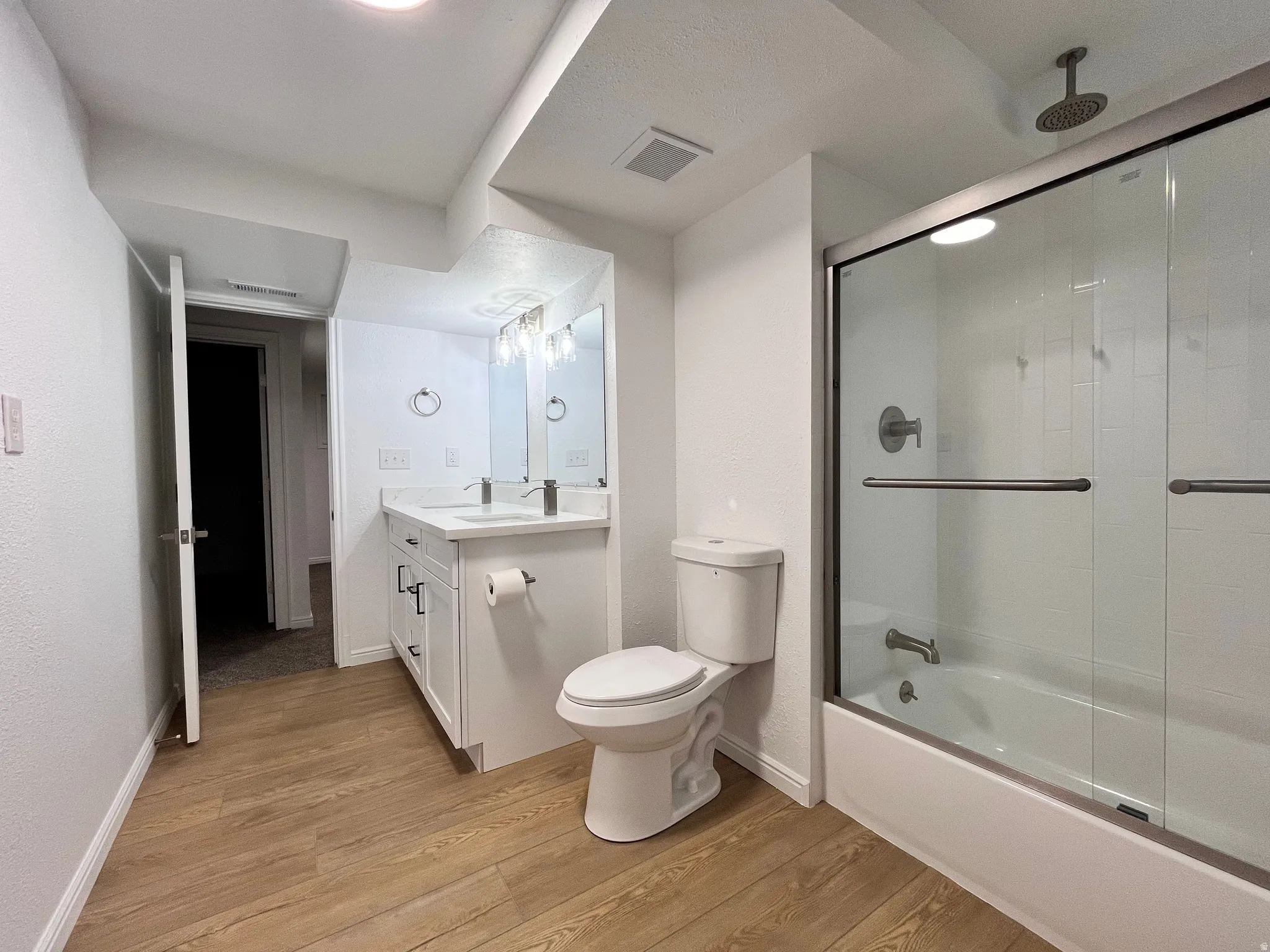 Bathroom featuring a dual vanity with white cabinetry, a glass-enclosed tub/shower combination with subway tile surround, wood-finish flooring, and recessed lighting