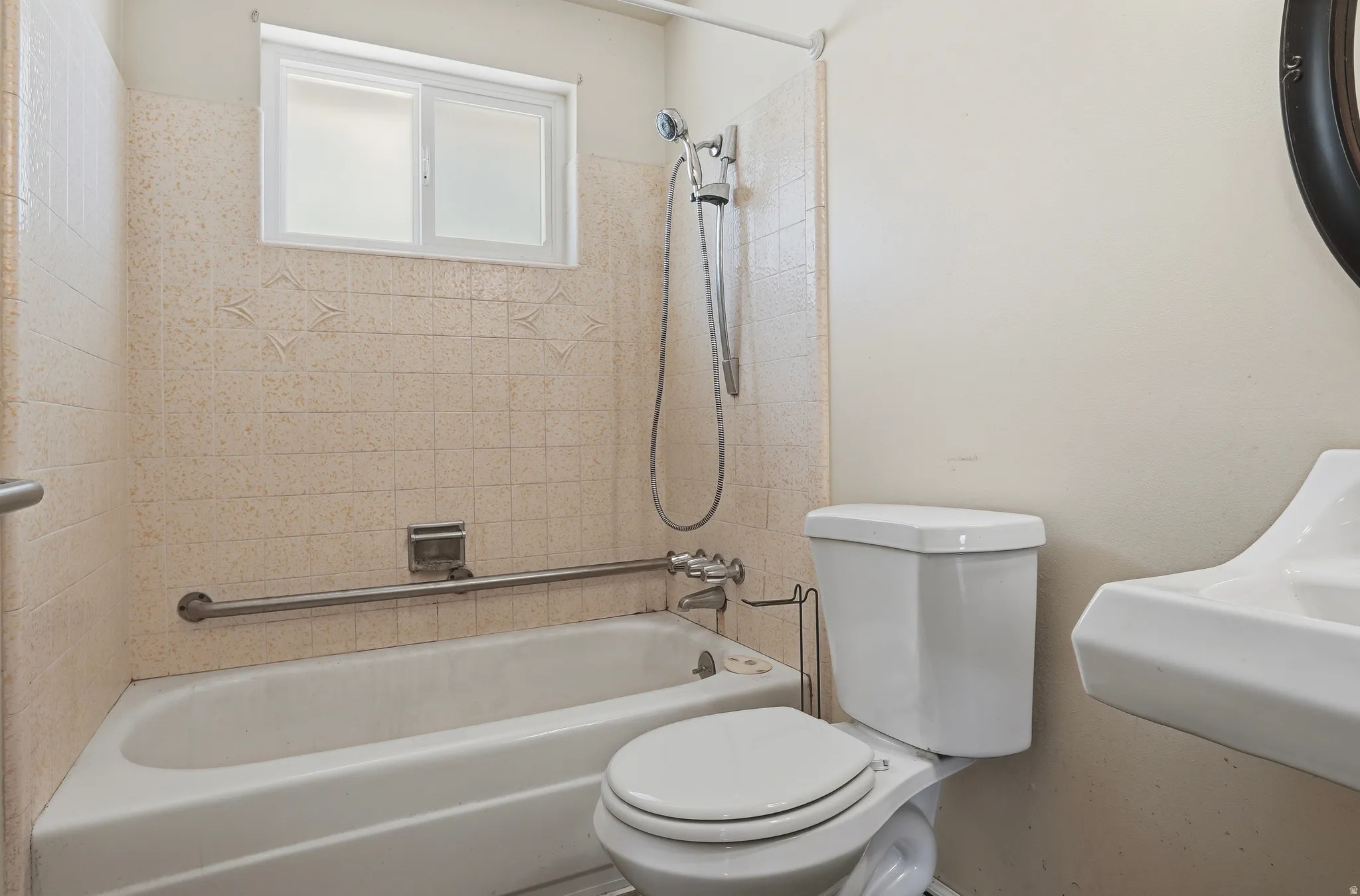 Full bathroom featuring a white bathtub with a shower head, light-toned wall tile, a window with frosted panes, and a white toilet