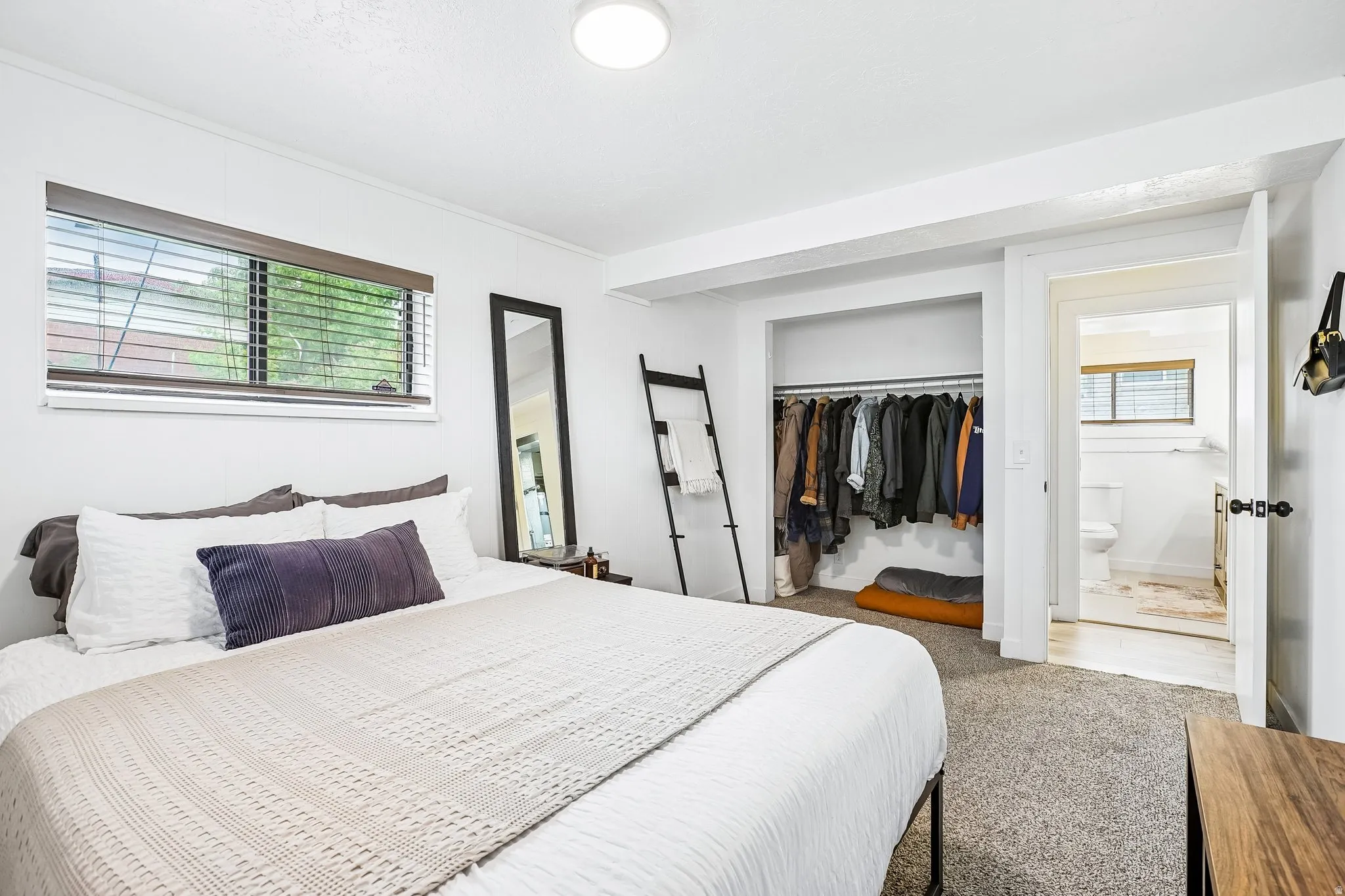 Bedroom featuring a window with blinds, recessed ceiling lighting, wall-to-wall carpeting, an open closet with clothing rod, and an adjacent bathroom with a window and a toilet