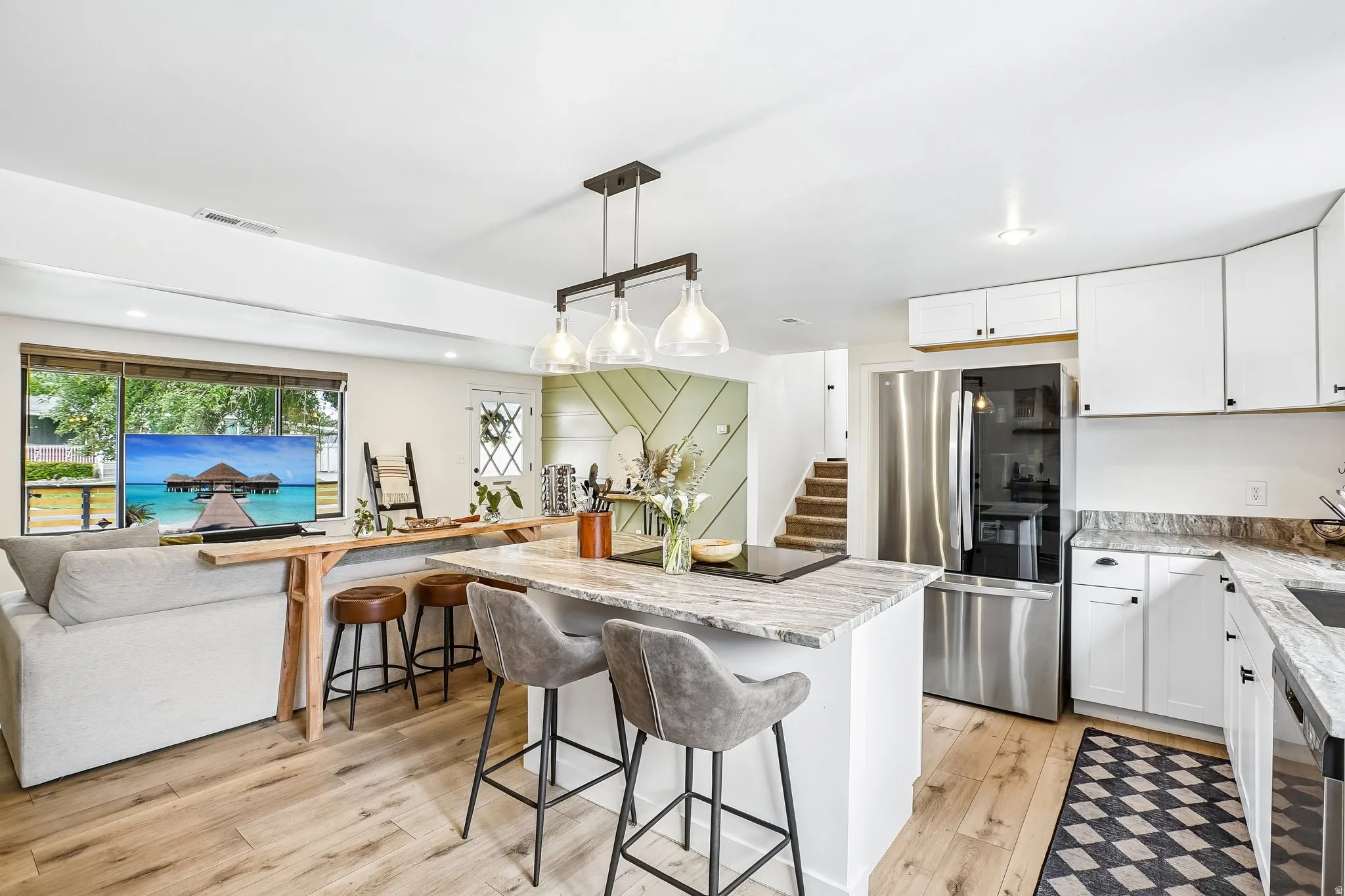 Open-concept living space featuring wood-finish flooring, a kitchen island with stone countertop, white cabinetry with black hardware, stainless steel refrigerator, and large window