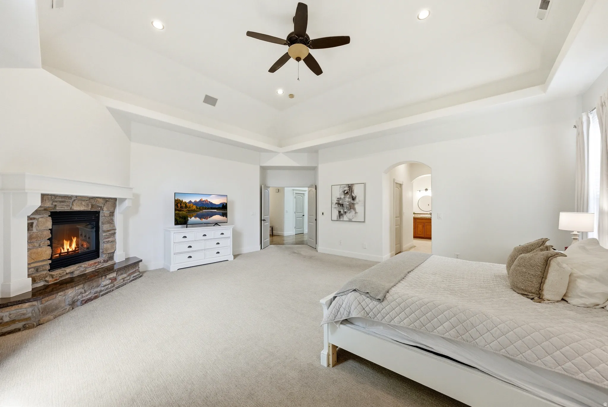 Spacious bedroom featuring a stone fireplace with a white mantel, tray ceiling, recessed lighting, and a ceiling fan