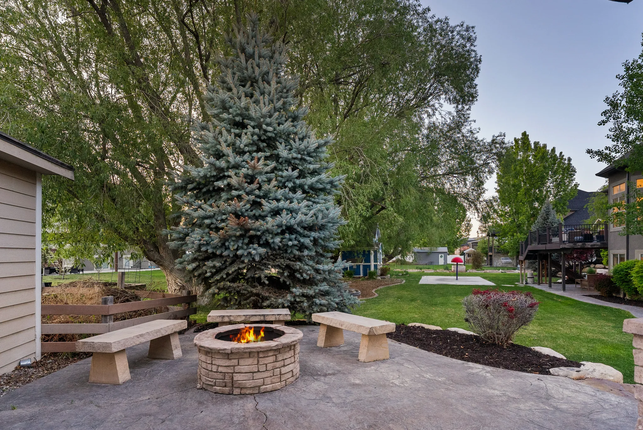 Stamped concrete patio with a stone fire pit and three stone benches, surrounded by a manicured lawn, mature trees, and landscaping