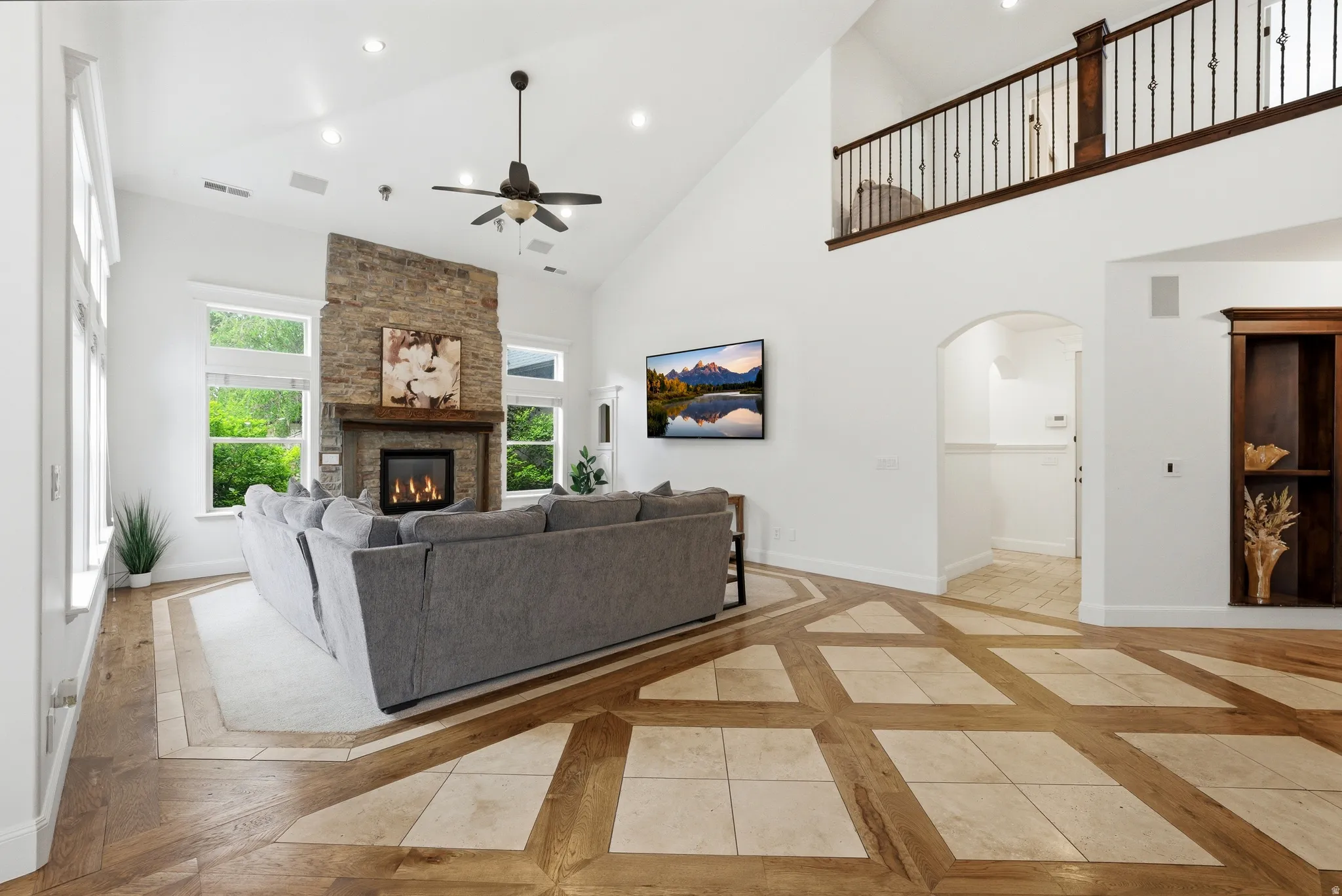 Vaulted living room featuring a stone fireplace with gas insert, large windows, custom wood-finish flooring with tile accents, and an upper-level balcony with wrought iron railing