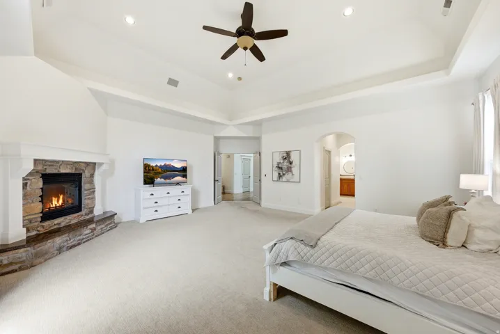 Spacious bedroom featuring a stone fireplace with a white mantel, tray ceiling, recessed lighting, and a ceiling fan
