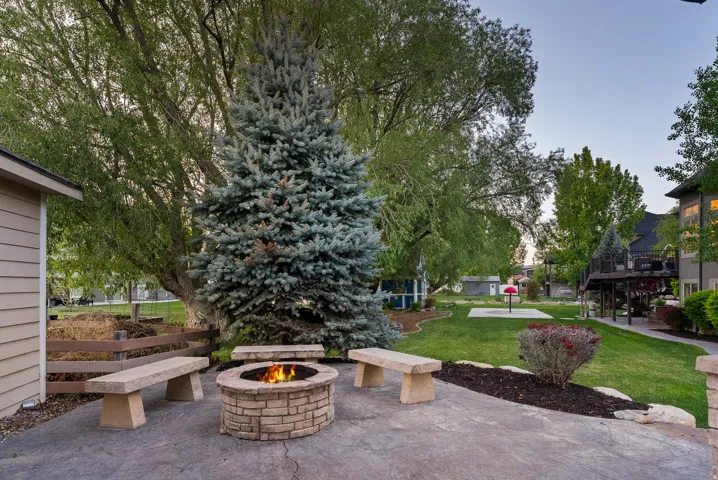 Stamped concrete patio with a stone fire pit and three stone benches, surrounded by a manicured lawn, mature trees, and landscaping