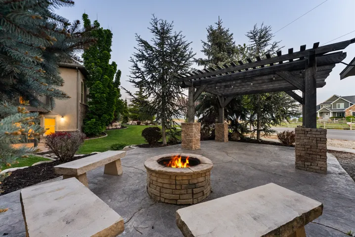 Stamped concrete patio featuring a stone-clad fire pit, built-in stone benches, a dark-stained wood pergola with stone-clad pillars, and surrounding evergreen trees
