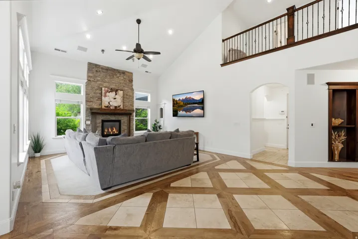 Vaulted living room featuring a stone fireplace with gas insert, large windows, custom wood-finish flooring with tile accents, and an upper-level balcony with wrought iron railing