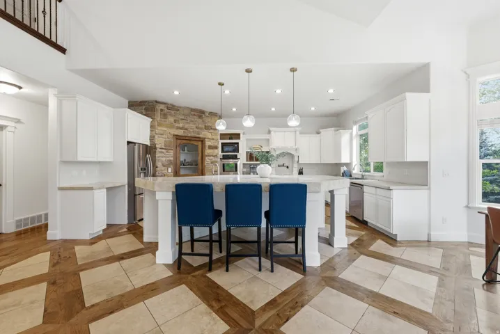 Spacious kitchen featuring an oversized island with seating, stainless steel appliances, abundant white cabinetry, a natural stone accent wall, and unique wood-finish flooring with tile inserts