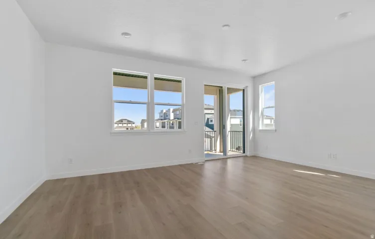 Spacious room featuring wood-finish flooring, recessed lighting, white walls, multiple windows, and a glass sliding door opening to a balcony with a black metal railing