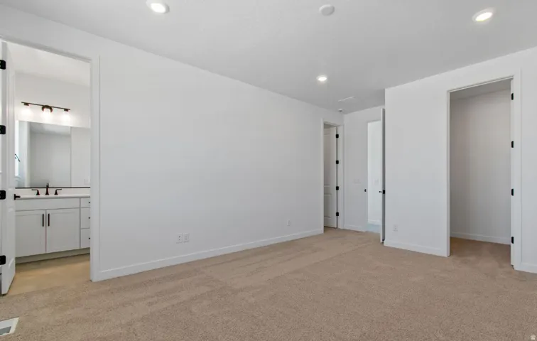 Spacious room featuring neutral carpeting, white walls, recessed lighting, and an open doorway leading to a bathroom with a white vanity and dark fixtures