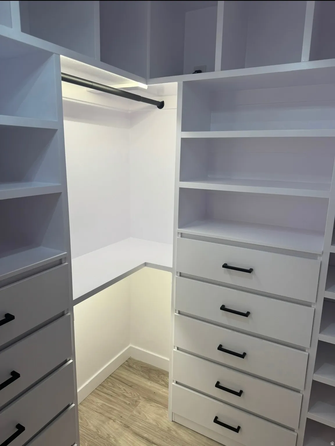 White built-in closet system featuring multiple shelving units, integrated drawers with dark hardware, a clothes hanging rod, and under-shelf lighting