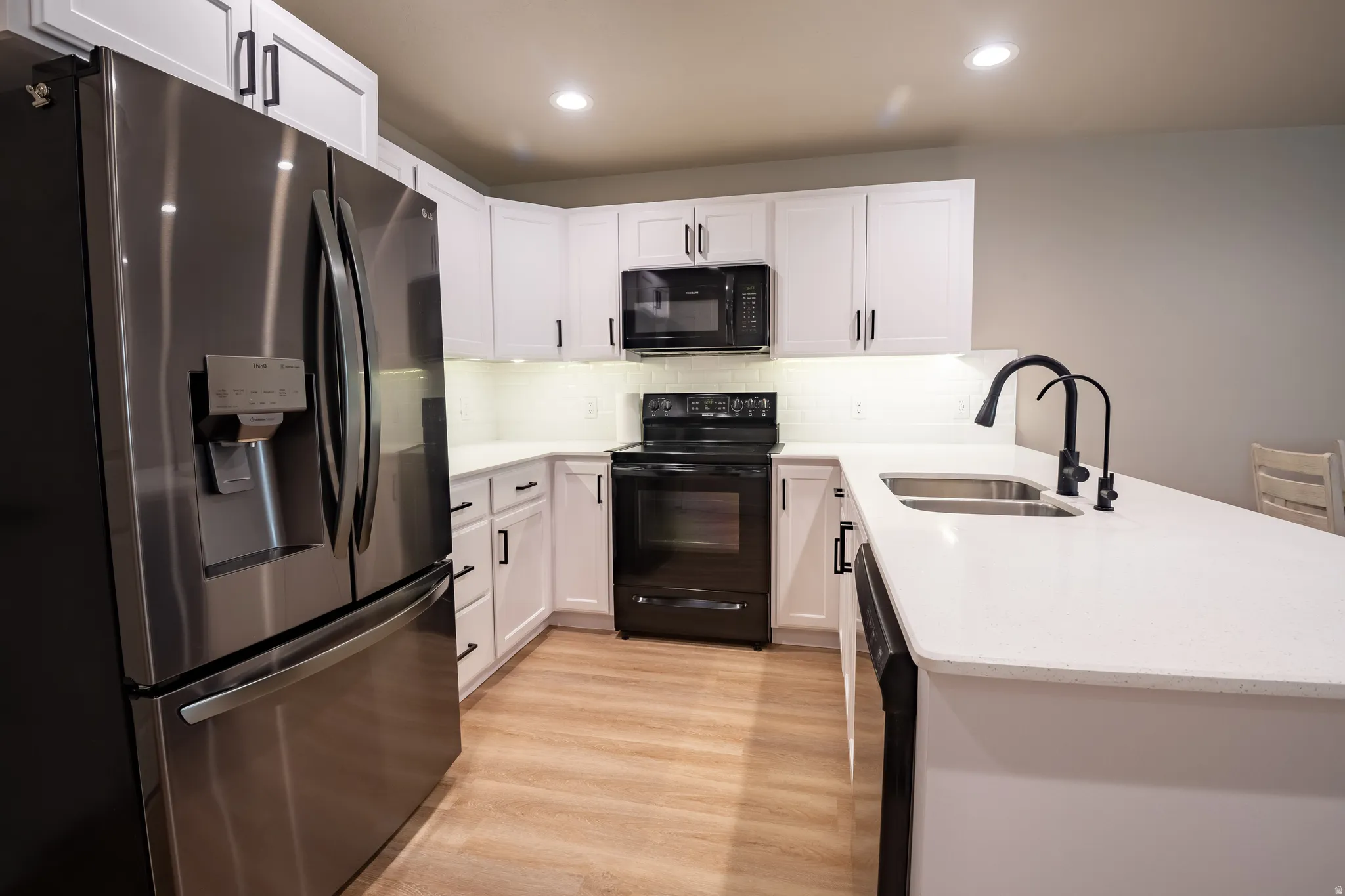 Kitchen featuring white cabinetry, light-toned wood-finish flooring, stainless steel French door refrigerator, black range, and white countertops with an undermount double-basin sink and matte black faucet