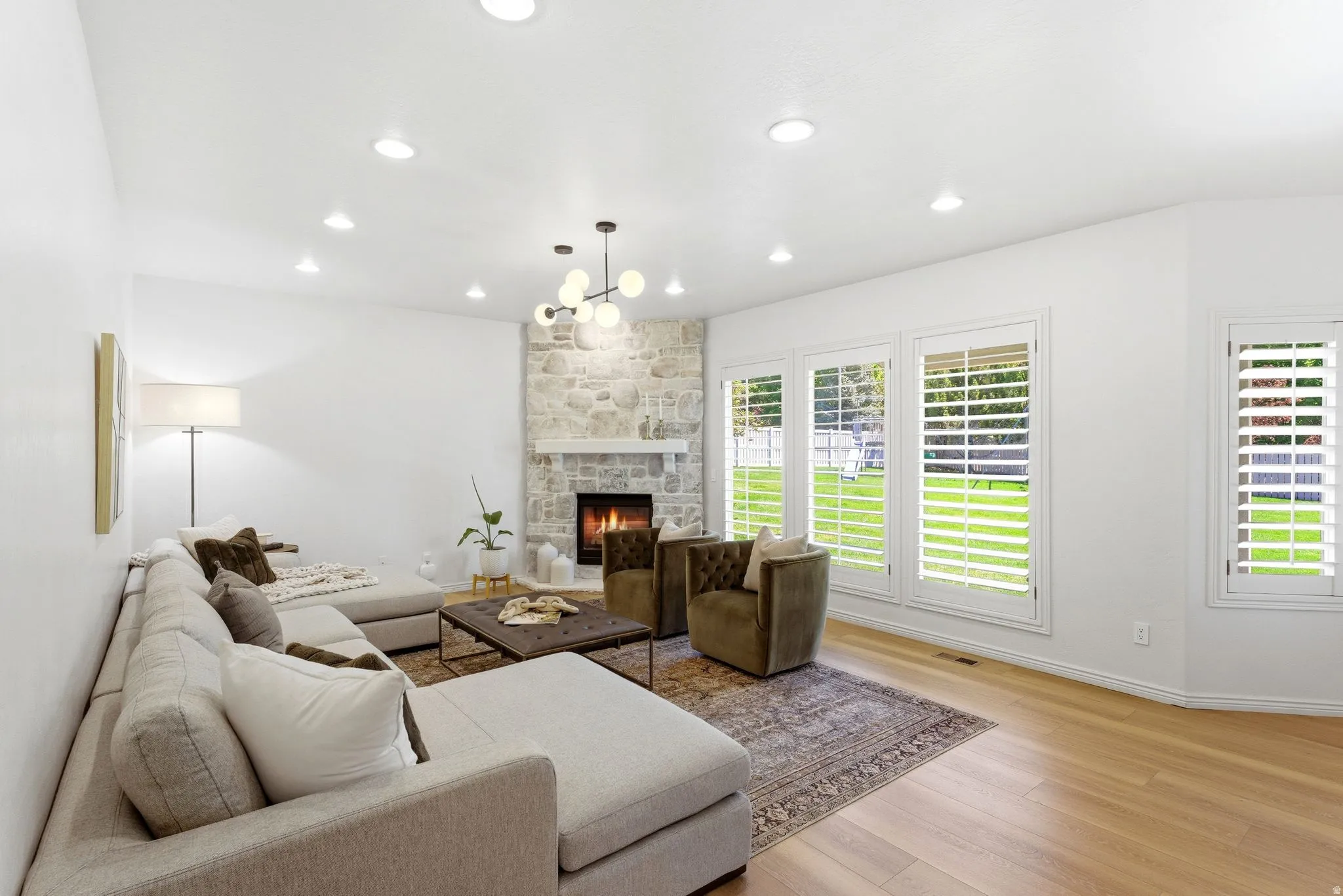 Spacious living area featuring a stone fireplace with a white mantel, light wood-finish flooring, recessed lighting, and multiple windows with white plantation shutters