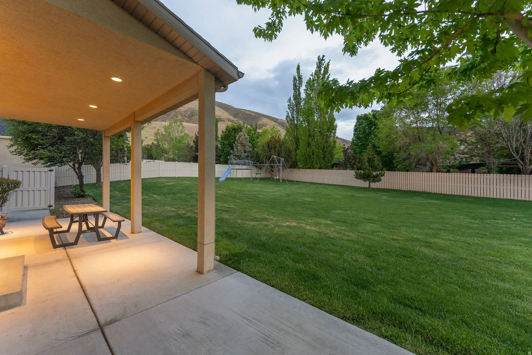 Expansive backyard featuring a covered patio with recessed lighting, concrete flooring, a lush green lawn, and a light-toned privacy fence