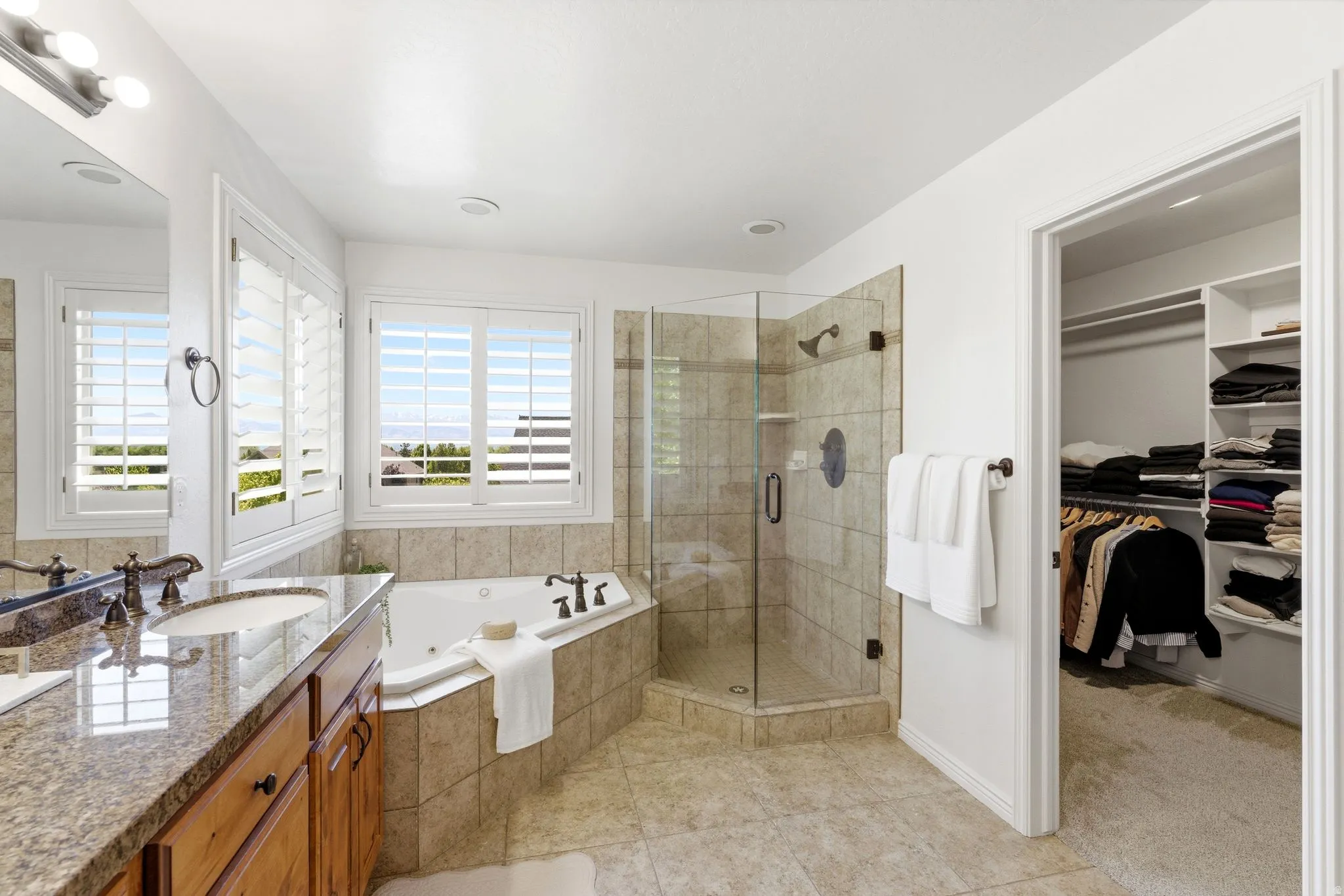 Bathroom featuring a corner soaking tub, a separate glass-enclosed shower with tile surround, a double vanity with granite countertop and wood cabinetry, and a walk-in closet with built-in shelving