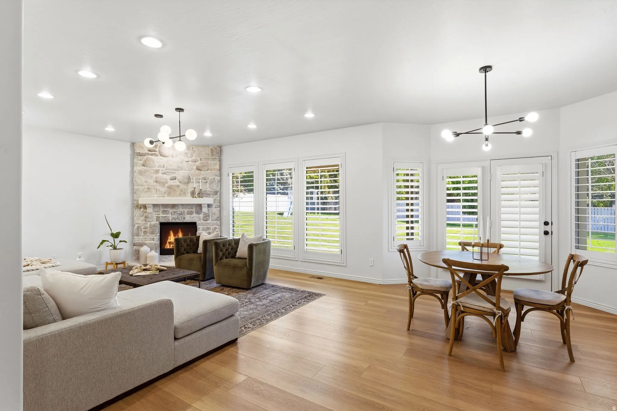 Bright living area featuring wood-finish flooring, a stone-clad fireplace with a white mantel, and multiple windows with plantation shutters