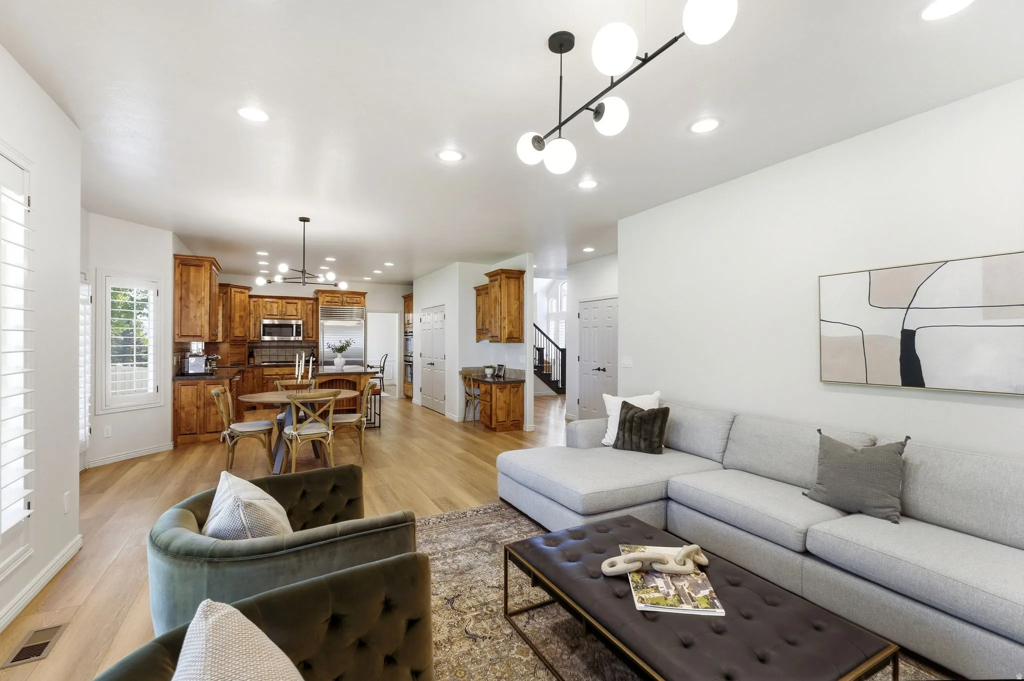 Open-concept living area featuring light wood-finish flooring, recessed lighting, white plantation shutters, and a kitchen with warm wood cabinetry