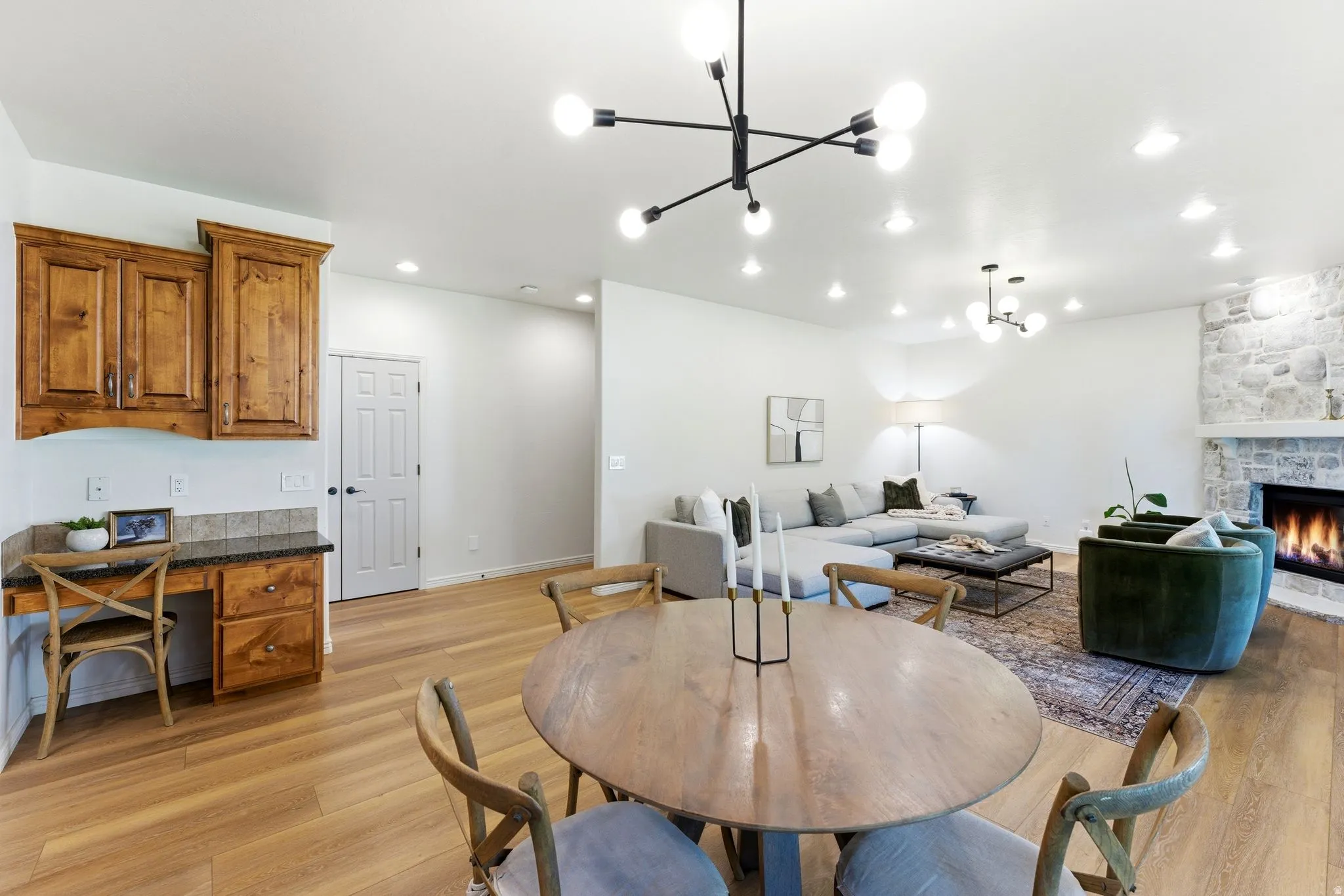 Open-concept living area featuring light wood-finish flooring, a stone fireplace with a white mantel, and a modern chandelier