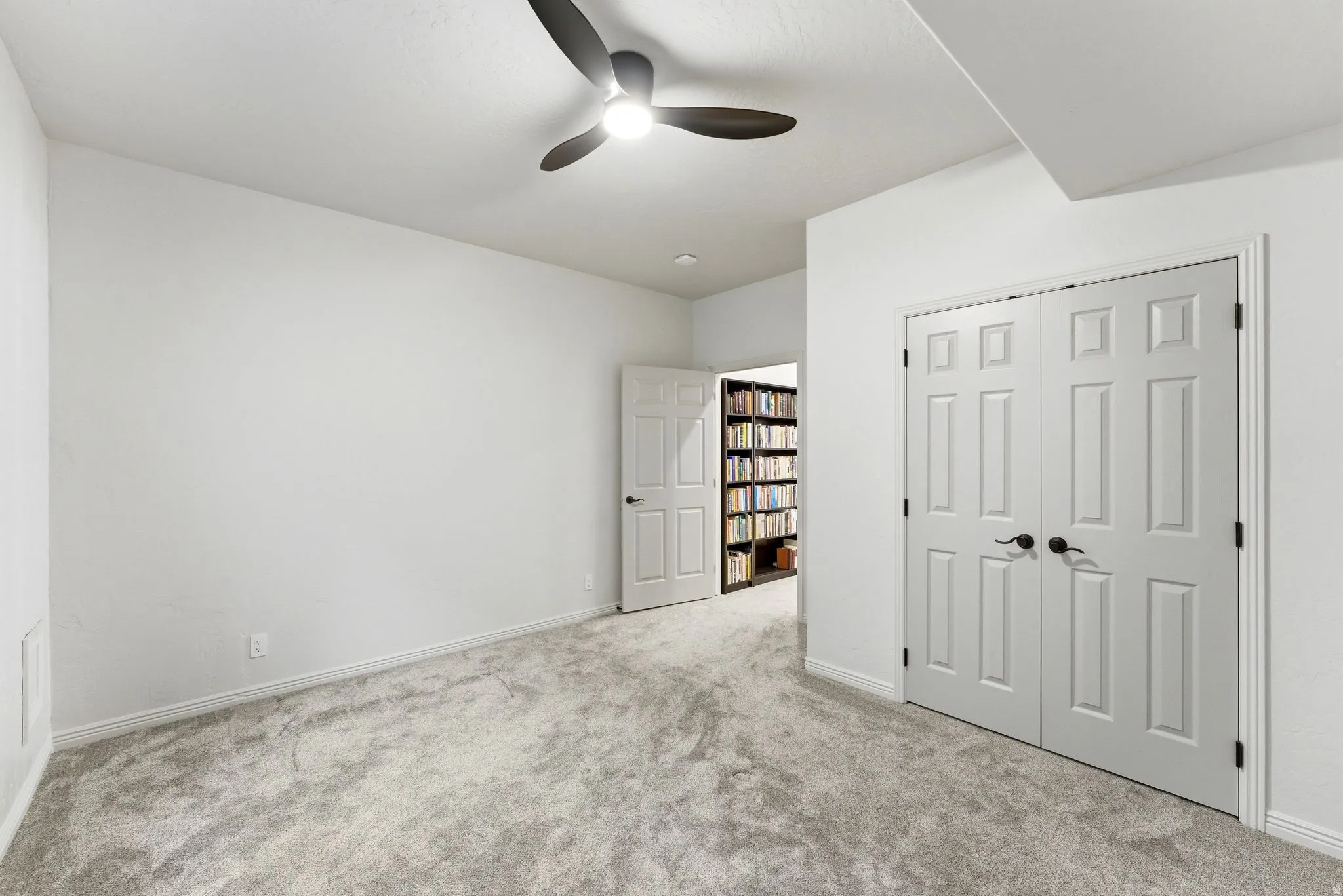 Room featuring light gray patterned carpet, white walls, a ceiling fan with integrated lighting, and white six-panel bifold doors with black hardware