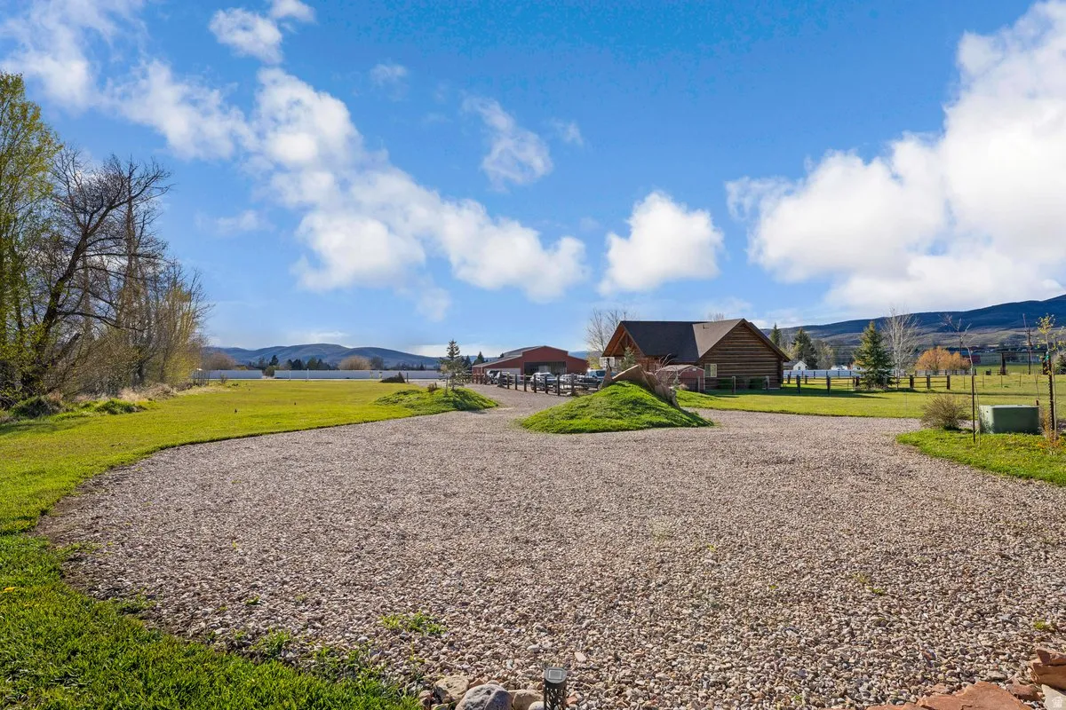 Gravel driveway leading to a rustic-style home with a gabled roof and natural wood siding, surrounded by expansive green lawns, mature trees, and distant mountains