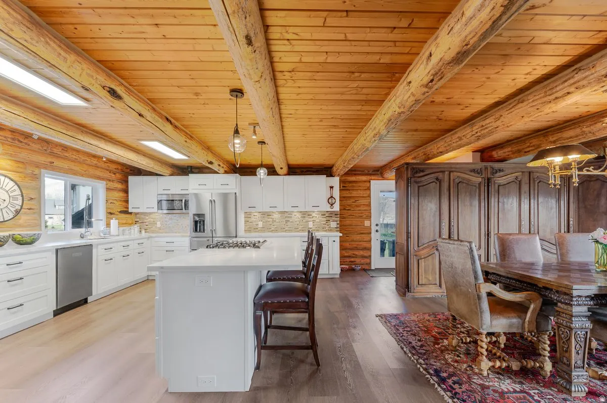 Open-concept kitchen and dining area featuring a wood-finish ceiling, exposed log beams, white cabinetry, a large kitchen island with counter seating, and stainless steel appliances