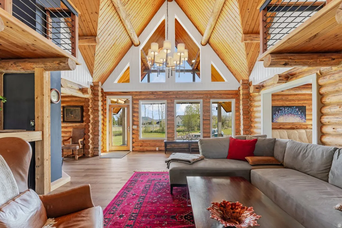Soaring cathedral ceiling with exposed log beams, wood-finish flooring, multiple windows providing natural light, and a second-story interior balcony with metal railings