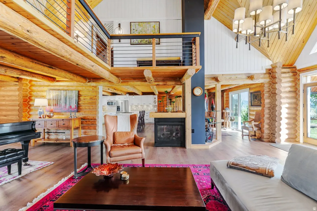 Two-story open-concept interior featuring prominent log walls and exposed timber beams, wood-finish flooring, a contemporary fireplace, and a second-story balcony with metal railing