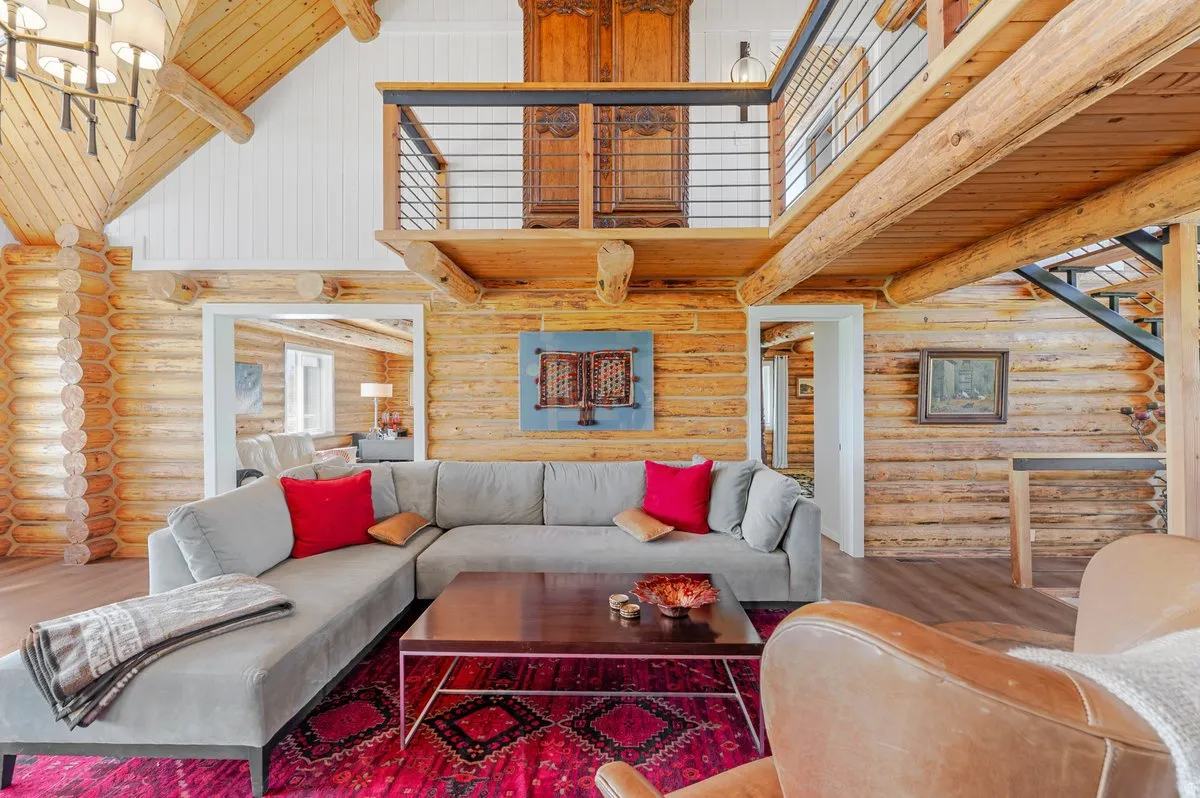 Living area showcasing log construction, exposed wood beams, a mezzanine balcony with metal railing, vaulted white plank ceiling, and wood-finish flooring
