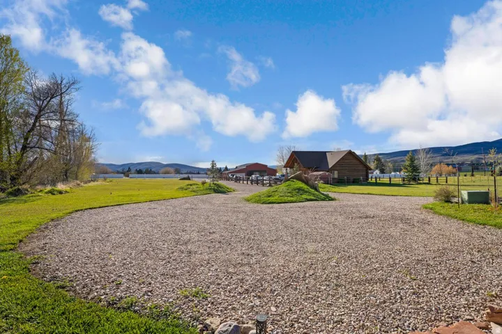 Gravel driveway leading to a rustic-style home with a gabled roof and natural wood siding, surrounded by expansive green lawns, mature trees, and distant mountains