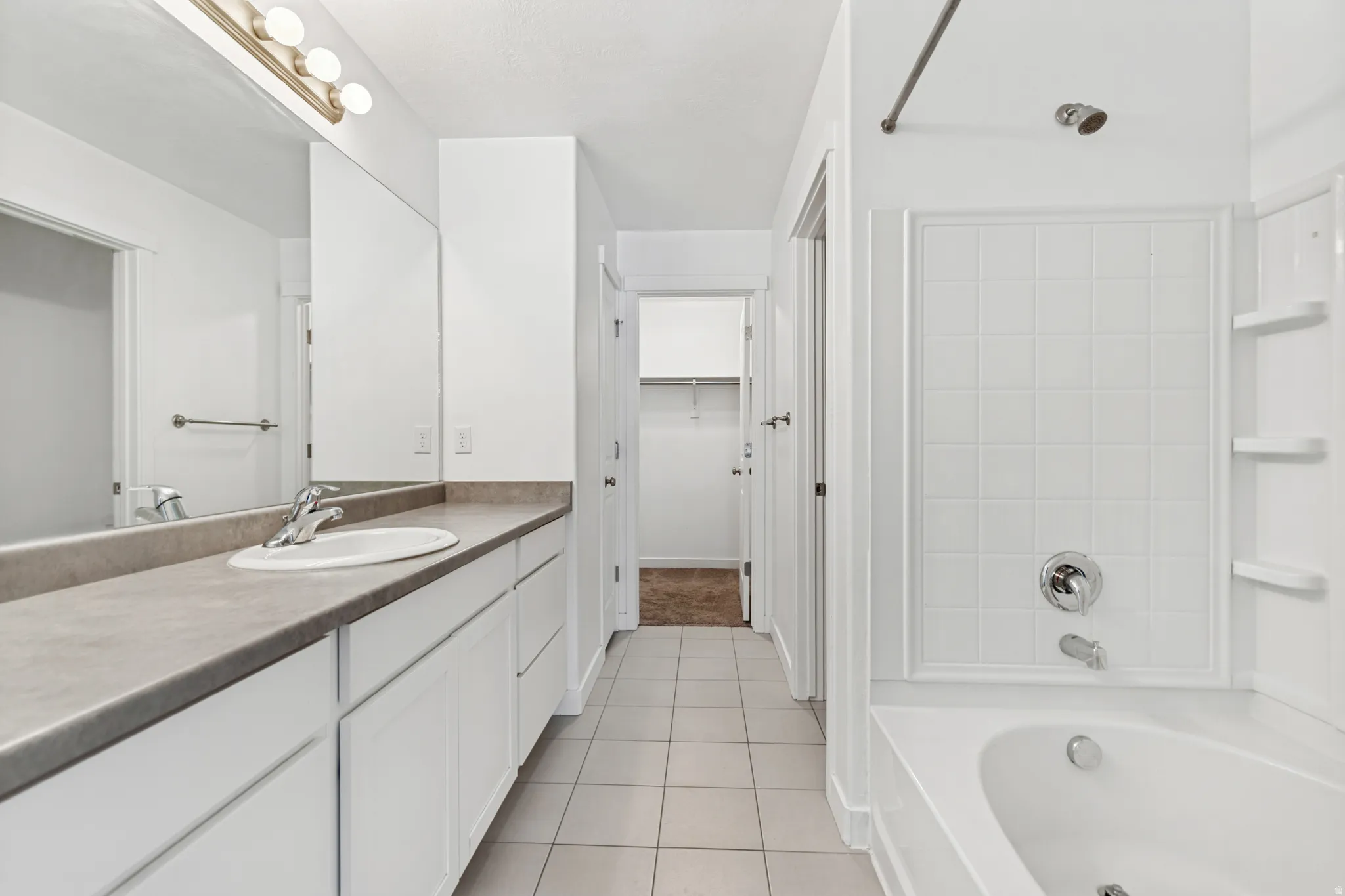 Bathroom featuring a white tile floor, an elongated vanity with a single basin sink and gray countertop, a large wall-mounted mirror with overhead lighting, and a bathtub with white tiled surround and built-in shelving