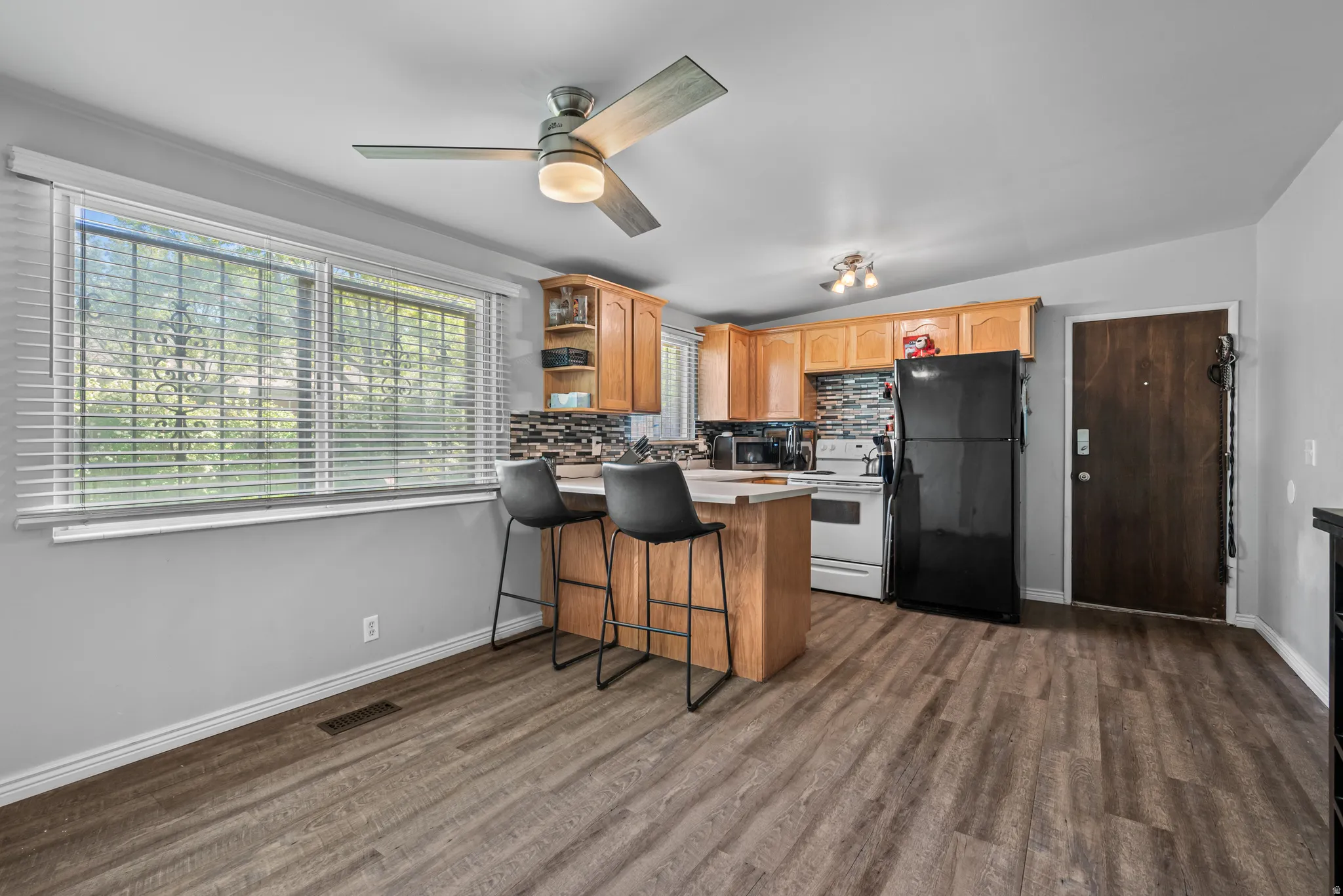 Kitchen and dining area featuring wood-finish flooring, light wood cabinetry, white appliances, and a mosaic tile backsplash