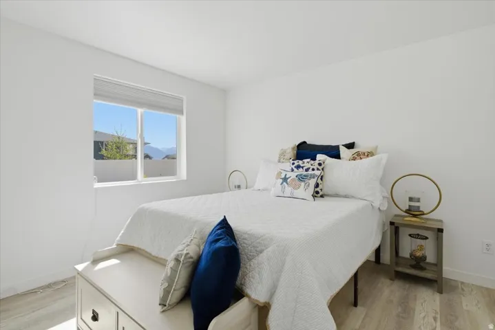 Bright bedroom featuring white walls, a large window with roller shades, and wood-finish flooring