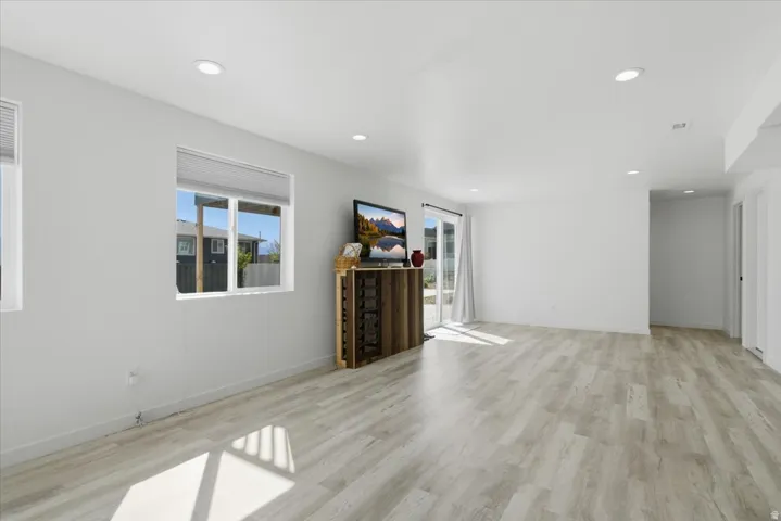 Spacious living area featuring light wood-finish flooring, recessed lighting, white walls, multiple windows with blinds, and a glass-paneled exterior door