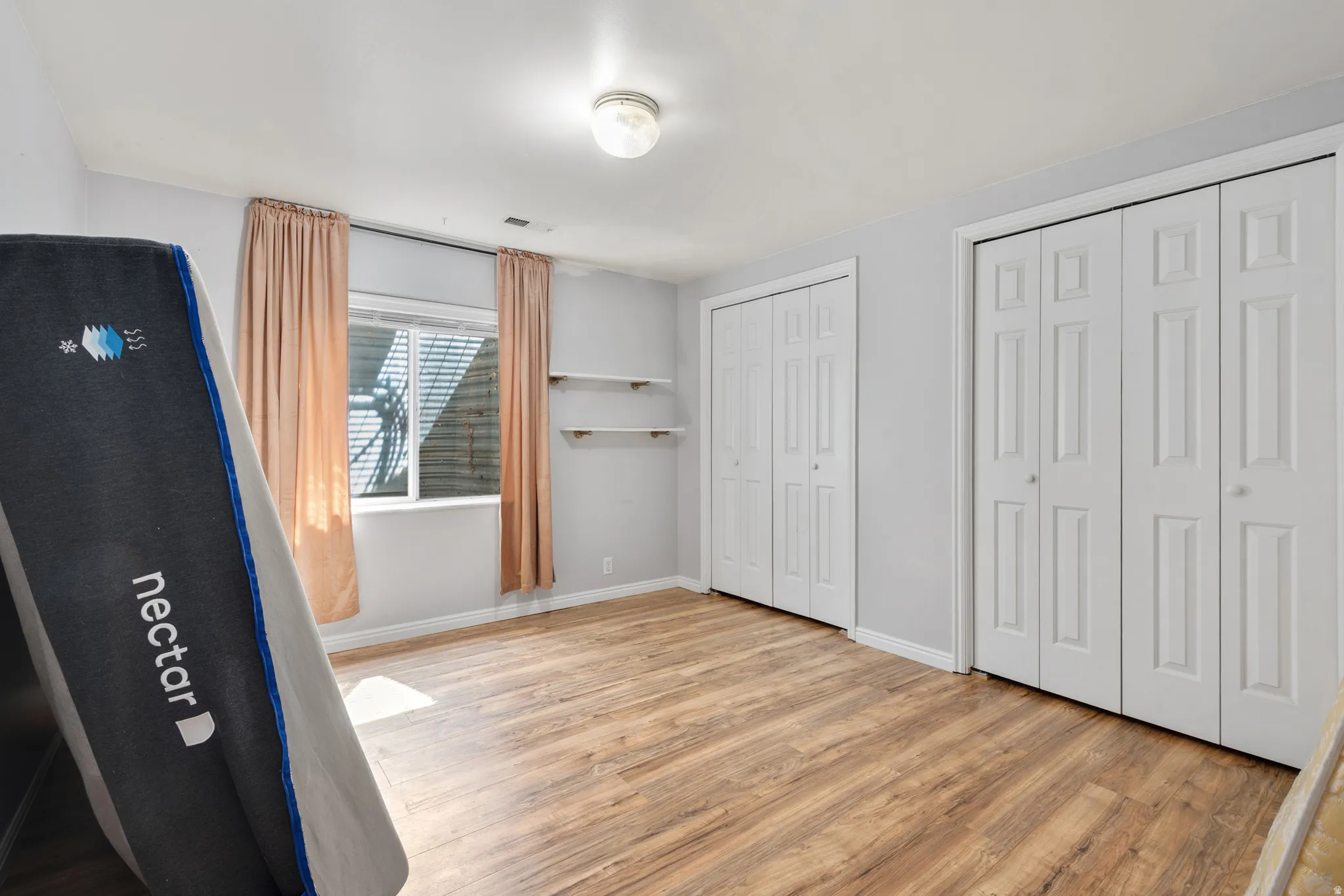 Room featuring wood-finish flooring, light grey walls, a window with horizontal blinds, and two sets of bi-fold closet doors
