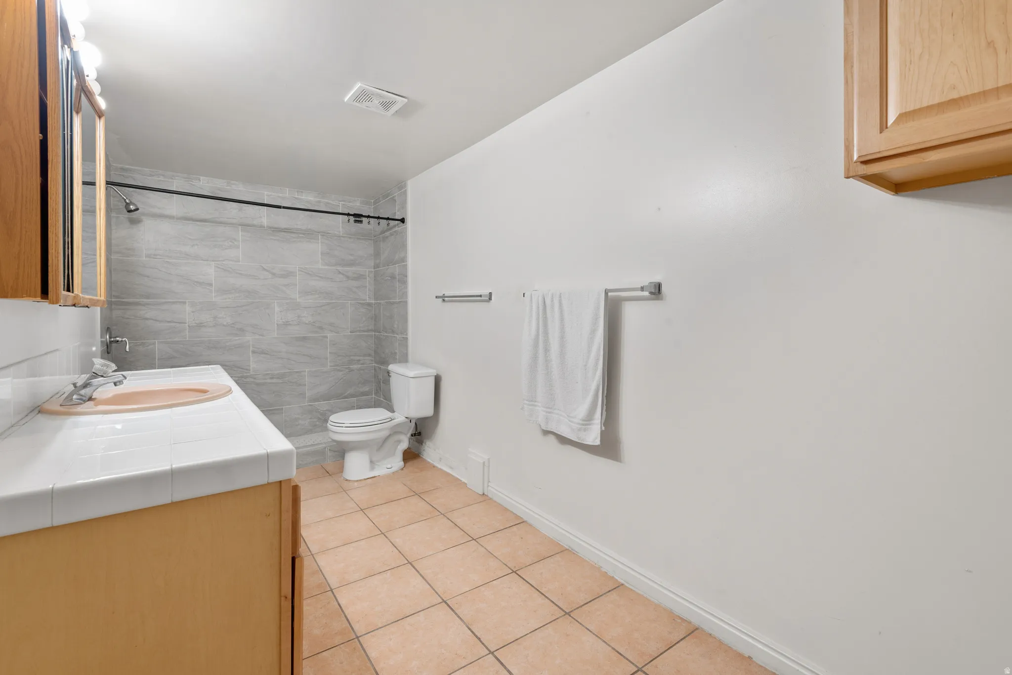 Bathroom featuring light gray tiled shower surround, a white tiled vanity countertop, and a wood-finish vanity cabinet with integrated sink