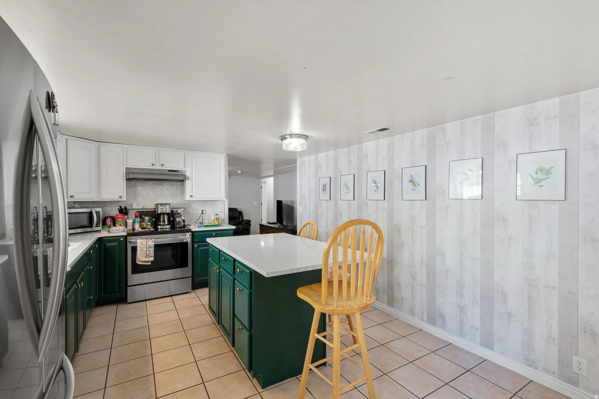 Kitchen featuring an island with a white countertop and dark green cabinetry, stainless steel appliances, two-tone cabinetry, a tiled floor, and a decorative paneled accent wall