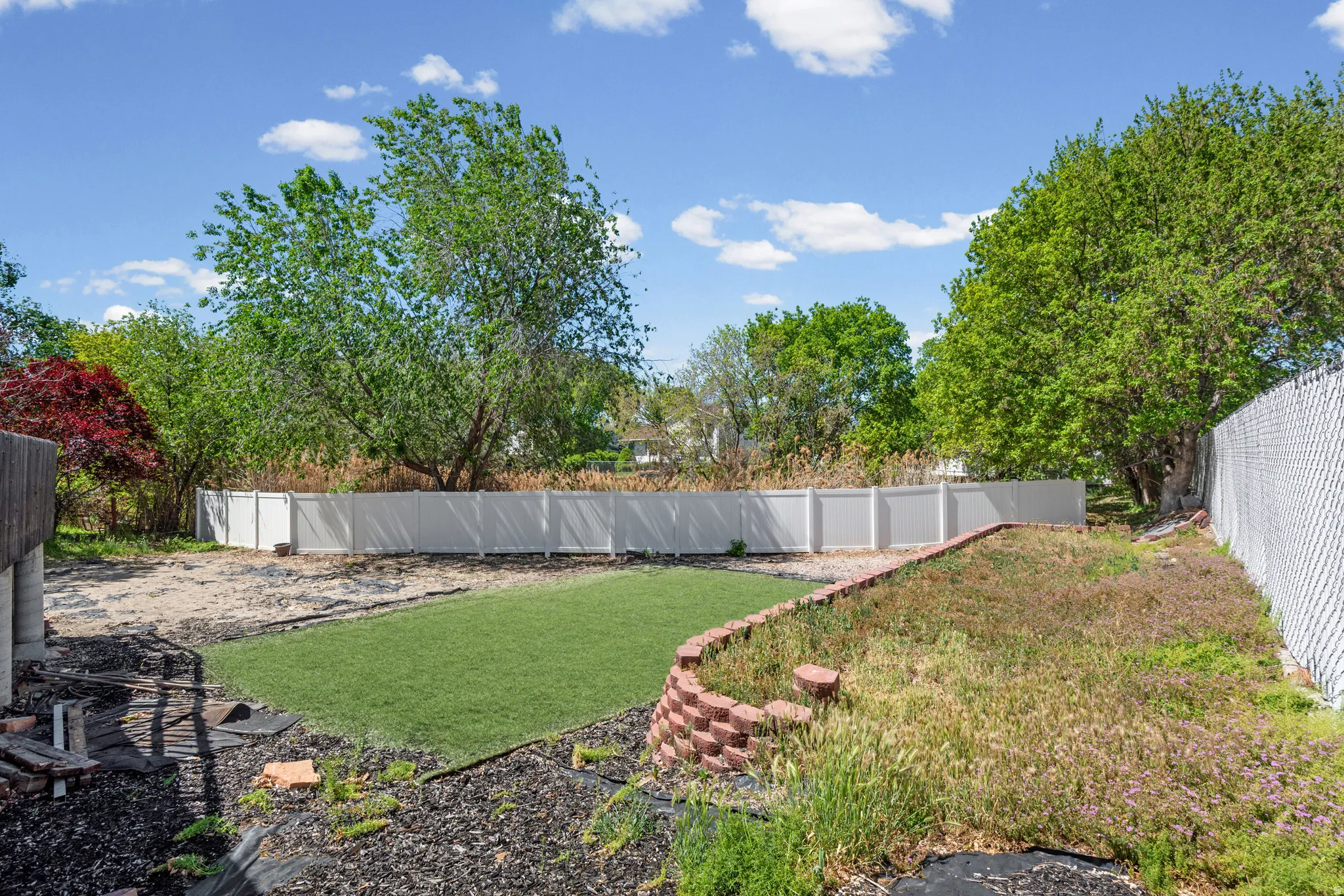 Expansive outdoor space featuring a white privacy fence, mature trees, a chain-link fence, and a terraced garden area with brick-style retaining walls
