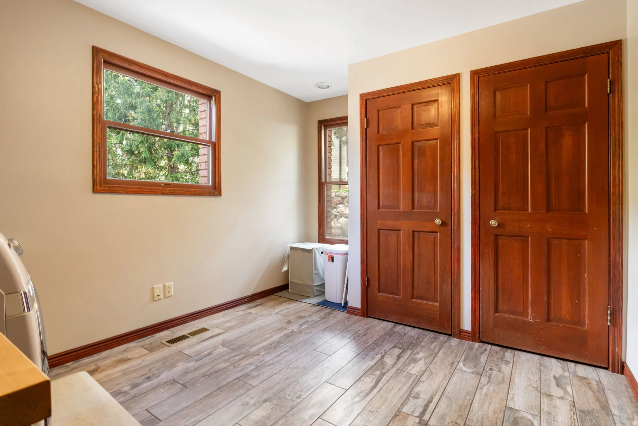 Room featuring light-toned walls, wood-finish flooring, two windows with wood trim, and two six-panel wood doors