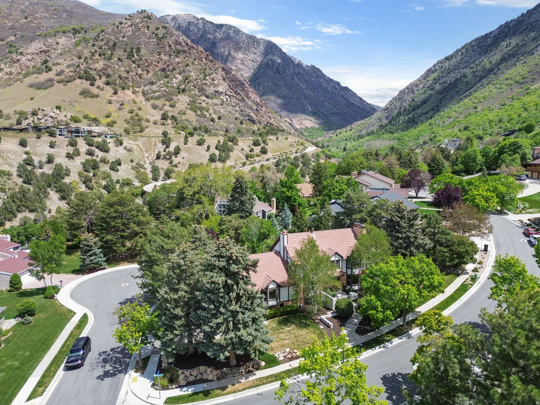 Residential property situated in a mountainous valley, featuring mature trees, a red tile roof, and a paved street with sidewalks