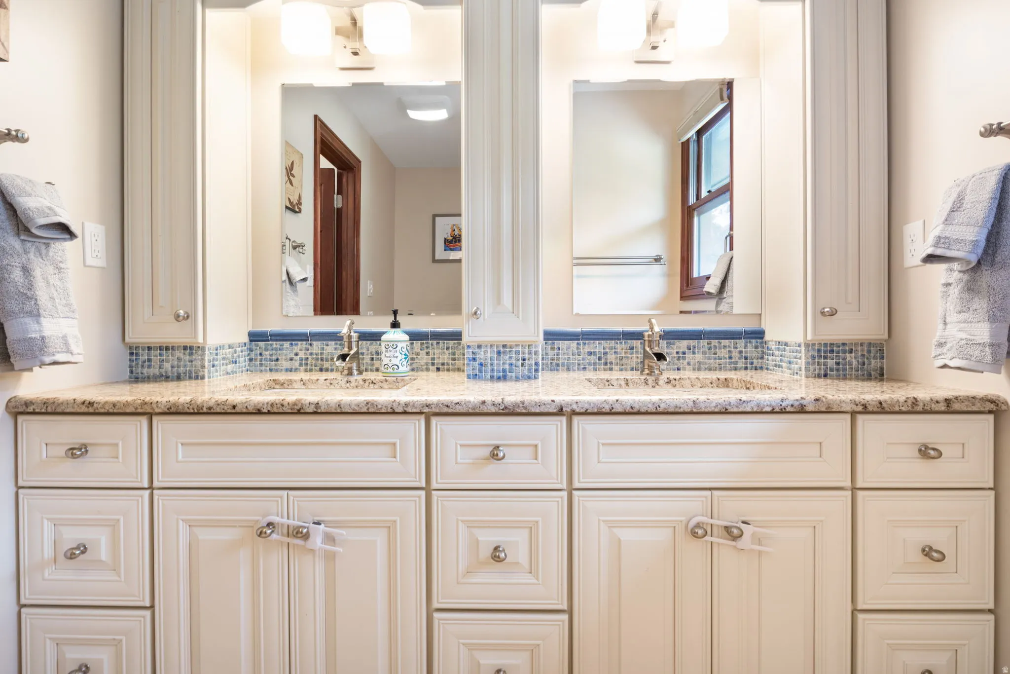 Double vanity featuring a light speckled countertop, dual undermount sinks with chrome faucets, a mosaic tile backsplash, and two framed mirrors with overhead lighting